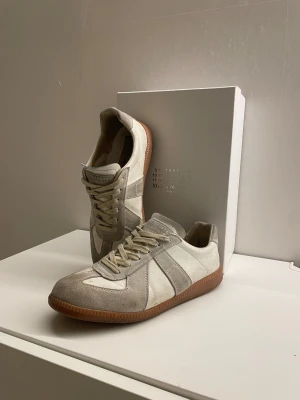 Maison Margiela gats - Maison Margiela sneakers i vitt med klassisk design. Skorna har snörning, rund tå och platt gummisula i brunt. Ovandelen är i mocka och skinn, med subtila paneler och logga på tungan. Perfekt för dig som gillar stilrena och trendiga sneakers.