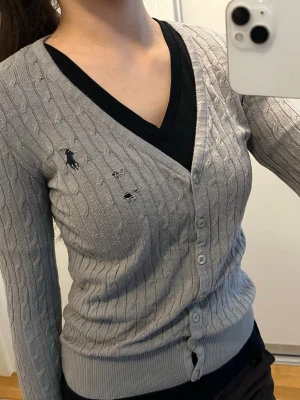 Grå kabelstickad kofta från Ralph Lauren - Snygg grå kabelstickad kofta från Ralph Lauren med V-ringning och knäppning framtill. Klassisk broderad logga på bröstet och ribbade muddar vid ärmslut och nederkant. Perfekt att slänga över en t-shirt eller topp för en clean look. Har defekter vid loggan