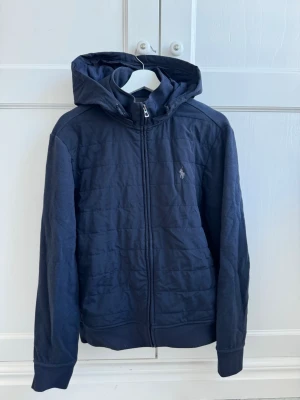 Mörkblå hoodiejacka från Polo Ralph Lauren - Mörkblå quiltad hoodiejacka från Polo Ralph Lauren med dragkedja framtill och broderad logga på bröstet. Jackan har huva, ribbade muddar och är tillverkad i ett mjukt material som ger en avslappnad look.