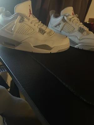 Vita Air Jordan 4 Retro sneakers - Säljer ett par vita Air Jordan 4 Retro sneakers med grå och röda detaljer. Skorna har klassisk meshpanel, synlig Air-sula och Jumpman-logga på häl och plös. Tillverkade i läder och textil för en clean och sportig look. Använda och smutsiga 