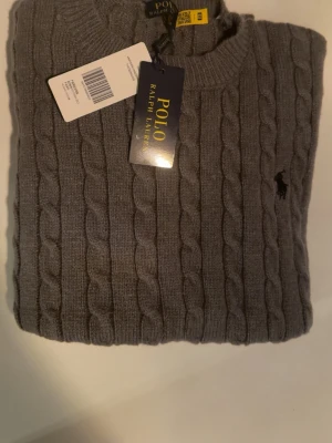 Grå kabelstickad tröja Polo Ralph Lauren - Kabelstickad grå tröja från Polo Ralph Lauren med rund halsringning och diskret broderad logga på bröstet. Klassisk modell med ribbade muddar vid ärmslut och nederkant. Perfekt för lager på lager och snygg till jeans.