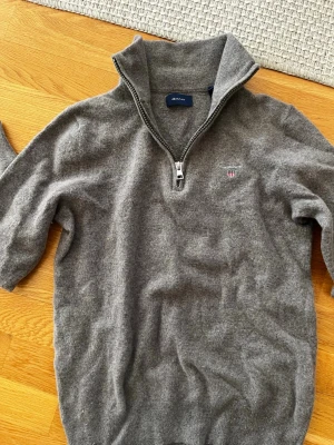 Grå stickad halvzip-tröja från Gant - Mysig brun stickad tröja från Gant med halv dragkedja och hög krage. Liten broderad logga på bröstet. Perfekt för lager-på-lager och passar till det mesta. Ribbstickade muddar vid ärmslut och nederkant. Den har krympt så passar xs / S i storlek. 