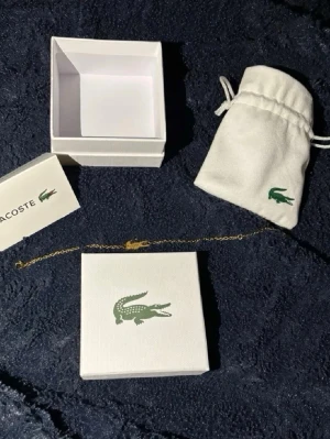 Guldfärgat armband från Lacoste - Snyggt och stilrent armband från Lacoste i guldfärgad metall. Armbandet har en tunn kedja och ett ikoniskt krokodil-hänge i mitten. Perfekt accessoar för att lyfta din outfit med en touch av lyx och sportig känsla. Levereras med originalask och tygpåse. Båda två för 600kr  annars 1 för 500kr en manlig armband och en kvinnlig armband 