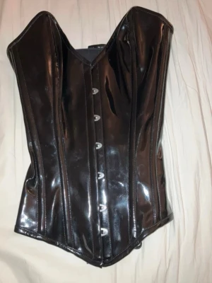 Svart blank korsett – Corsets Island – XS - Supersnygg svart korsett i blank finish från Corsets Island. Storlek XS.  • Snörning baktill (justerbar passform) • Hyskor framtill • Formande och stabil modell • Ger en jättefin siluett 🖤  Perfekt för goth/alt-look, fest, layering över skjorta eller klänning – eller som statement piece.  I fint skick. Inga skador, dragkedjor eller hyskor fungerar som de ska.  Pris: 500 kr (kan diskuteras vid snabb affär)