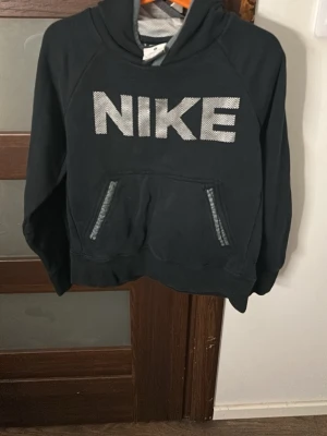 Svart Nike hoodie med stor logga - Svart hoodie från Nike med stor vit logga på bröstet. Klassisk känguruficka framtill, ribbade muddar och huva med grått innerfoder. Perfekt för en sportig och avslappnad look.
