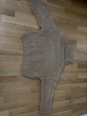 Beige/brun kabelstickad polotröja - Säljer en beige/brun kabelstickad polotröja med breda ribbade muddar vid ärmslut och nederkant. Tröjan har ett klassiskt flätmönster på framsida samt ärmar. Perfekt för dig som gillar en mysig och trendig look.