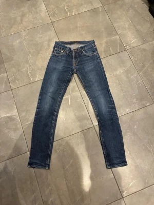 Blå slim jeans från Nudie Jeans - Säljer ett par klassiska blå jeans från Nudie Jeans med slim passform och fem fickor. Jeansen har snygga slitningar och kontrastsömmar samt detaljerade bakfickor med Nudie-jeans loggan. Passar perfekt till dig som är 150-155
