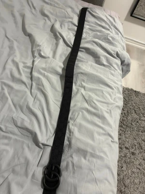 Svart bälte med GG-spänne från Gucci - Snyggt svart bälte från Gucci med det klassiska GG-spännet i matt svart metall. Bältet har diskret mönster med GG-loggor längs hela bandet och är tillverkat i skinn. Perfekt för att lyfta vilken outfit som helst.