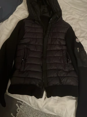 Svart pufferjacka från Moncler - Snygg svart pufferjacka från Moncler med quiltade partier framtill och huva. Jackan har dragkedja, två fickor med knappstängning och Moncler-logga på ärmen. Ärmar och rygg är i stickad kvalitet för extra komfort.