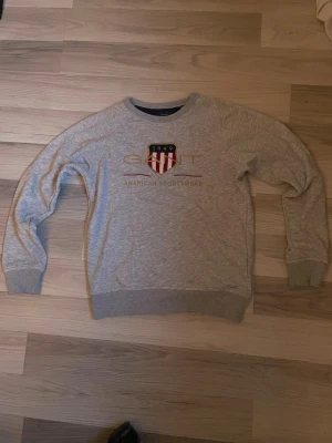 Grå sweatshirt från Gant - Snygg grå sweatshirt från Gant med broderad logga och texten 'American Sportswear' framtill. Passar folk som är 145-154cm långa. Tröjan har rund halsringning, ribbade muddar och klassisk passform. Perfekt för en avslappnad och sportig stil. Tröjan har inga större skador och är i bra skick.