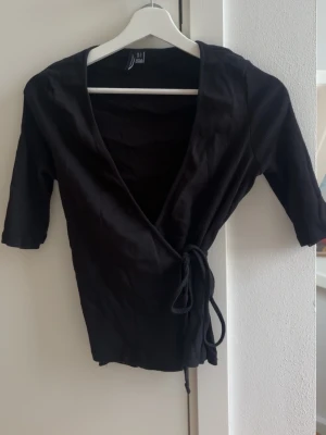 Svart omlottopp med knyt - Snygg svart omlottopp från vero moda med knyt i sidan och djup v-ringning. Toppen har halvlånga ärmar och är tillverkad i mjuk bomull, perfekt för en trendig och enkel look. Jätte fint att ha ett spets linne eller ett vanligt linne under! Lite defekter där man trär snöret igenom men inget som syns alls! 