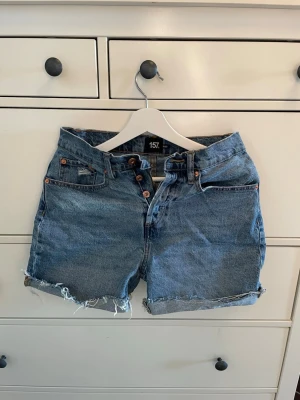Blå denimshorts från 157 - Säljer ett par klassiska blå denimshorts från 157 med hög midja och råa, fransiga benslut. Shortsen har fem fickor, knappgylf och bälteshällor. Perfekta för sommaren och ger en avslappnad vibe.