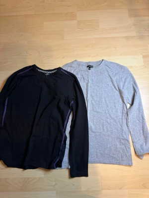 2 långärmade tröjor, KappAhl & svart - Två långärmade tröjor, en grå från KappAhl i mjuk bomull och en svart med lila sömmar och rund hals. Perfekta basplagg till garderoben, båda har enkel och clean design. Passar till jeans eller mjukisbyxor.