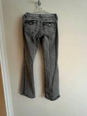 Grå bootcut jeans med fickdetaljer - Snygga grå jeans med bootcut-modell och dekorativa fickor bak. Jeansen har klassisk femficksdesign och är tillverkade i ett mjukt denimtyg. Perfekta för dig som gillar en avslappnad men trendig stil.