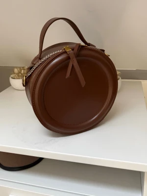 Rund brun handväska i läder - Snygg rund handväska i brunt läder med guldfärgade detaljer och kedja. Väskan har ett praktiskt handtag upptill och dragkedja med läderflärp. Perfekt för dig som vill ha en unik och trendig accessoar.