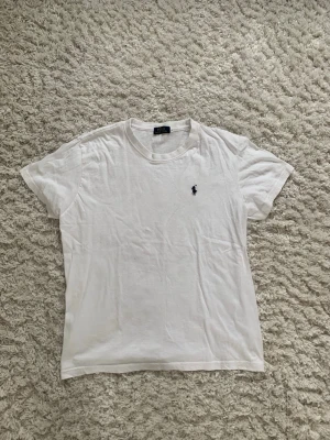 Vit t-shirt från Ralph Lauren - Klassisk vit t-shirt från Ralph Lauren med den ikoniska broderade loggan på bröstet. T-shirten har rund halsringning och korta ärmar. Tillverkad i mjuk bomull för en skön och avslappnad stil.