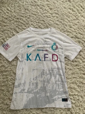 Vit Al Nassr matchtröja från Nike - Säljer en vit Al Nassr matchtröja från Nike med klubbmärke och sponsorloggor i blått och lila. Tröjan har korta ärmar, rund hals och subtilt mönster i tyget. Perfekt för fotbollsträning eller att bära som supporter. Materialet är lätt och andas bra.