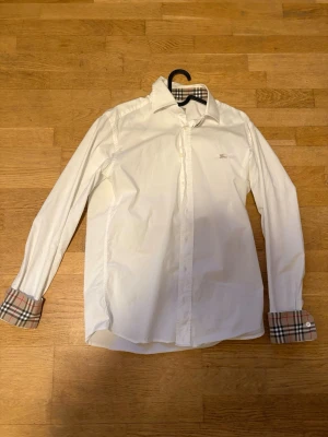 Vit Burberry skjorta med rutiga detaljer - Klassisk vit skjorta från Burberry med ikoniska beige, röda och svarta rutiga detaljer på krage och manschetter. Skjortan har lång ärm, knappar framtill och är tillverkad i bomull. Perfekt för dig som vill ha en stilren look med en twist.