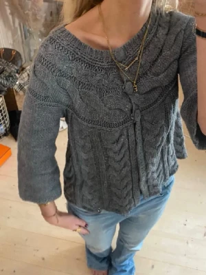 Grå kabelstickad kofta med knytband - Mysig grå kabelstickad kofta med 3/4-ärmar och knytband framtill. Koftan har ett brett mönster över bröstet och en avslappnad passform. Perfekt att slänga över en t-shirt eller linne för en chill look.