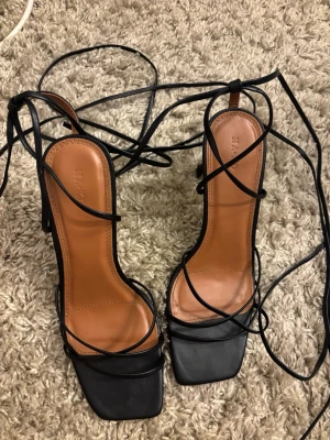 Svarta strappy pumps från NA-KD - Säljer ett par trendiga svarta pumps från NA-KD med tunna remmar och fyrkantig tå. Skorna har höga klackar och långa snörningar som kan lindas runt ankeln. Innersulan är brun och materialet ser ut att vara konstläder. Perfekta för en stilren look. Endast använda 1 gång på balen.. ena plastbiten på klacken har dock lossnat.