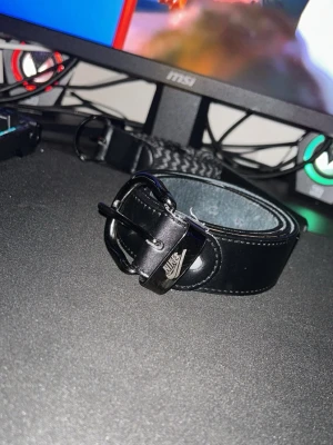 Svart skärp i skinn från Nike - Snyggt svart bälte från Nike i skinn med klassiskt spänne och diskret Nike-logga på metallspännet. Perfekt accessoar för att lyfta din outfit med en sportig touch. Passar till jeans eller byxor och ger en clean look.