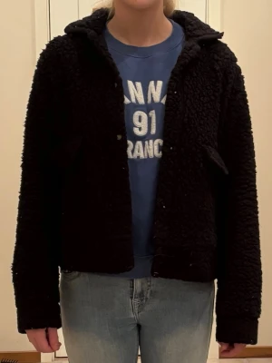 Svart teddyjacka med huva - Mysig svart teddyjacka med huva och knappar framtill. Jackan har två snedställda fickor och är tillverkad i fluffigt teddy-material som ger en cool och avslappnad look. Perfekt för dig som vill ha en trendig och bekväm jacka till vardags.