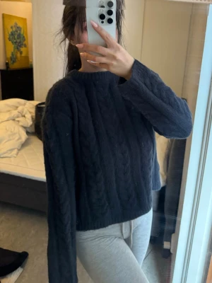 Mörkblå kabelstickad tröja - Mysig mörkblå kabelstickad tröja med croppad passform och långa ärmar. Tröjan har en rund halsringning och ett klassiskt flätmönster framtill. Perfekt för kyliga dagar och enkel att matcha med olika outfits.