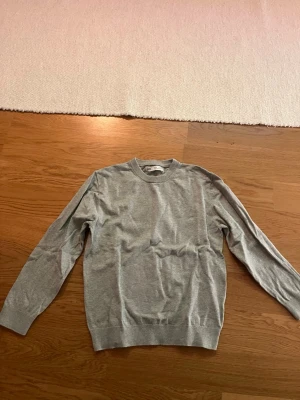 Grå basic tröja från ARKET - En stilren grå tröja från ARKET i mjukt bomullsmaterial. Klassisk rund halsringning och långa ärmar med ribbade muddar vid ärmslut och nederkant. Perfekt för en clean och enkel look. Dock så har den ett litet hål men det syns inte jätte mycket när man har på sig den