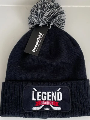 Mörkblå LEGEND Hockey mössa med tofs - Mörkblå ribbstickad mössa, LEGEND Hockey med broderad patch framtill. Mössan har en gråmelerad tofs på toppen och uppvikt kant. Perfekt för dig som gillar hockey och vill hålla dig varm. Eller bara du som vill va snyggast Pvg match. Från beechfield. 