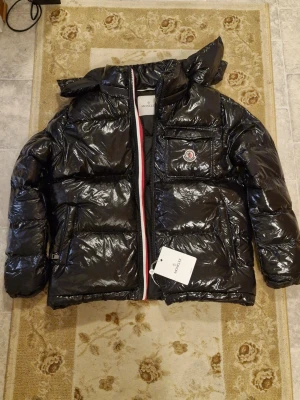 Svart glansig dunjacka från Moncler - Snygg svart dunjacka från Moncler med glansig finish och klassisk logga på bröstet. Jackan har huva, dragkedja med rödvit detalj och flera praktiska fickor. Perfekt för kalla vinterdagar och riktigt trendig stil.