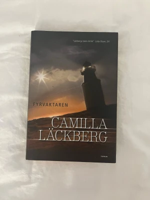 Fyrvaktaren av Camilla Läckberg📖 - Spännande kriminalroman av Camilla Läckberg, del sju i Fjällbackaserien. Omslaget är mörkt med motiv av en fyr mot dramatisk himmel. Boken är inbunden och har tydlig titel i vitt på framsidan. Perfekt för dig som gillar deckare och mysterier.