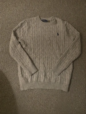 Beige kabelstickad tröja Polo Ralph Lauren - Kabelstickad beige tröja från Polo Ralph Lauren med rund halsringning och den klassiska mörkblå logotypen broderad på bröstet. Tröjan har långa ärmar och ribbade muddar vid ärmslut och nederkant. Perfekt för lager på lager och stilren vardagslook.