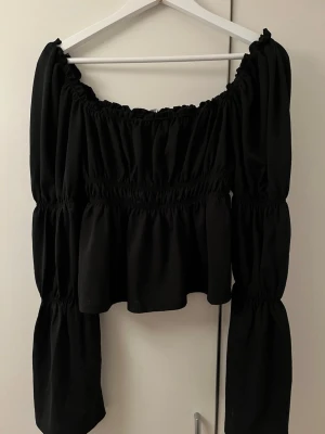 Svart offshoulder topp med volang från Gina tricot - Trendig svart offshoulder topp med långa, vida ärmar och volangdetaljer. Toppen har smock i midjan och runt axlarna för en snygg passform och croppad längd. Perfekt för dig som vill ha en statement piece i garderoben. Toppen går att bära som offshoulder eller som på bilden. Storlek M