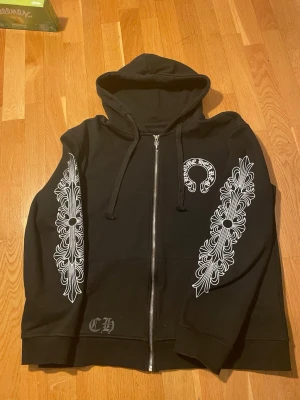 Svart Chrome Hearts hoodie med tryck - Säljer en svart hoodie från Chrome Hearts med dragkedja, huva och snörning. Hoodien har vita grafiska tryck längs båda ärmarna, logga på bröstet och stort tryck på ryggen. Materialet är mjukt och skönt bomullstyg. Perfekt för dig som gillar streetwear och unika detaljer.  Den är en S men passar om man är en M också
