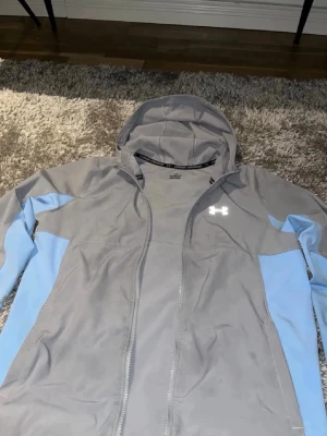 Grå och ljusblå vindjacka Under Armour - Snygg grå vindjacka från Under Armour med ljusblå detaljer på ärmarna. Jackan har huva, hel dragkedja och logga på bröstet. Perfekt för blåsiga dagar och sportiga outfits. Helt ny