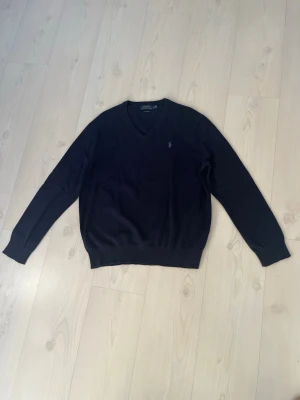 Mörkblå V-ringad tröja Polo Ralph Lauren - Klassisk mörkblå stickad tröja från Polo Ralph Lauren i mjuk pima bomull. Tröjan har V-ringning, långa ärmar och den ikoniska broderade loggan på bröstet. Perfekt att slänga på sig över en skjorta för en clean och stilren look.