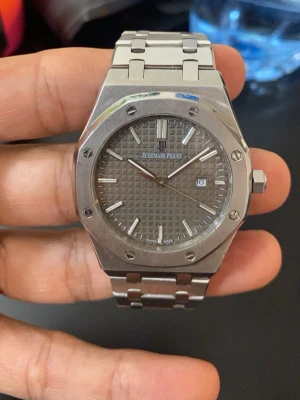 Audemars Piguet Royal Oak klocka kopi - Säljer en Audemars Piguet Royal Oak klocka i silverfärgat stål med ikonisk åttakantig boett och länkarmband. Urtavlan är grå med rutmönster och har datumvisning samt tydliga indexmarkeringar. En riktigt stilren och klassisk accessoar för dig som gillar exklusiva detaljer.