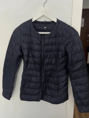 Svart dunväst från Uniqlo XS - Snygg svart dunväst från Uniqlo i storlek XS. Västen har rund halsringning, tryckknappar framtill och quiltad design. Tillverkad i lätt och smidigt material med dunfyllning som håller dig varm utan att kännas klumpig. Perfekt för lager-på-lager och enkel att matcha.