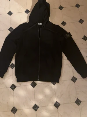 Svart hoodie från Stone Island - Säljer en svart hoodie från Stone Island med dragkedja framtill och klassisk logotyp-patch på vänster ärm. Hoodien har huva och ribbade muddar. Perfekt för dig som gillar streetwear och vill ha en stilren look.
