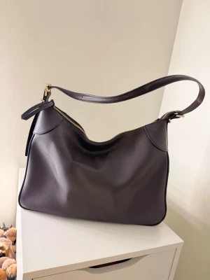 Mörkbrun axelväska i läderlook - Stilren mörkbrun axelväska i mjukt skinn med enkel, rundad form och guldfärgade metalldetaljer. Väskan har dragkedja upptill och bred axelrem för en bekväm look. Perfekt för dig som gillar minimalistisk och klassisk stil.