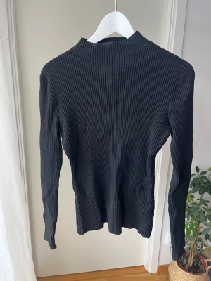 Svart ribbstickad tröja med hög krage - Säljer en svart ribbstickad tröja med lång ärm och hög krage. Tröjan har en smal passform och är perfekt för lager på lager. Materialet känns mjukt och stretchigt, vilket gör den bekväm att bära hela dagen.