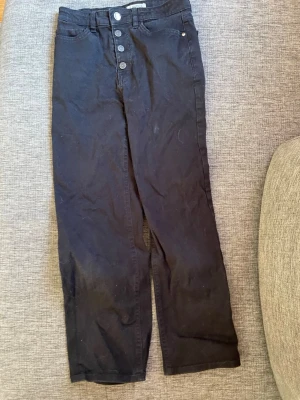 Svarta raka jeans med knappar - Säljer ett par svarta jeans med raka ben och hög midja. Jeansen har knappgylf med fyra synliga knappar och klassiska fem fickor. Materialet är denim i bomull. Perfekta till en avslappnad och trendig stil.