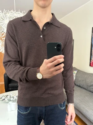 Brun långärmad pikétröja - Mörkbrun långärmad pikétröja med krage och knappar framtill. Tröjan har ribbade muddar vid ärmslut och nederkant, och är tillverkad i ett mjukt, tunt material som ger en stilren look. 