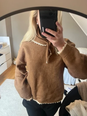 Brun stickad tröja med scarf - Mysig brun stickad tröja med knytband i halsen och dekorativ vit spetskant vid krage, ärmslut och nederkant. Tröjan har en avslappnad passform och långa ärmar, perfekt för kyligare dagar.