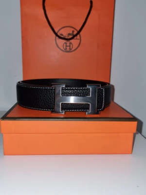 H bälte - Snyggt svart skärp från H med klassiskt Hformat metallspänne. Skärpet är gjort i exklusivt präglat skinn och levereras i den ikoniska orangea H boxen. Perfekt accessoar för att lyfta din outfit med en lyxig touch.