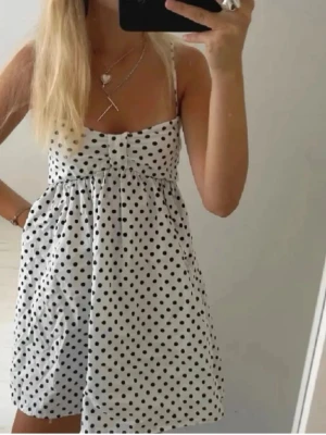Vit prickig kort klänning - Söt vit kort klänning med svarta prickar och tunna axelband. Klänningen har en lätt utställd passform och knappar framtill. Perfekt för varma dagar och ger en fräsch och ungdomlig look.