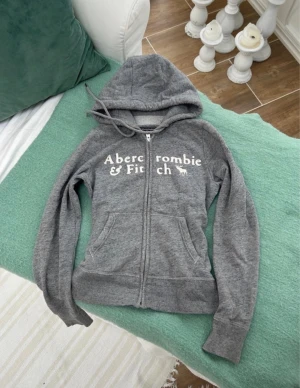 Grå hoodie från Abercrombie & Fitch - Snygg grå zip-hoodie från Abercrombie & Fitch med vit logga och älgtryck på bröstet. Hoodien har dragkedja, två fickor fram och justerbar huva med snören. Tillverkad i mjukt sweatshirtmaterial som är perfekt för chill dagar.