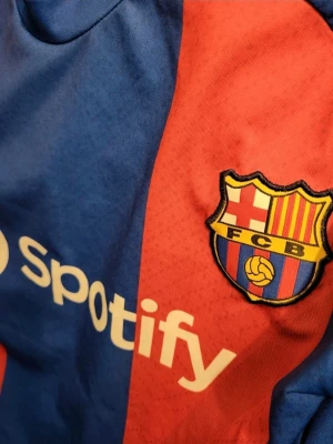 Nike FC Barcelona 2023/24 hemmastadion. Araújo storlek M - Säljer en officiell FC Barcelona matchtröja från Nike i blått och rött med klubbmärke och Spotify-logga på bröstet. Tröjan har korta ärmar, rund hals och är tillverkad i ett lätt, ventilerande funktionsmaterial. Perfekt för fotbollsträning eller att bära på matchdag.