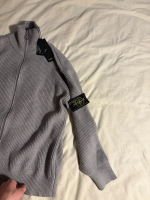 Ljusgrå stickad tröja Stone Island - Säljer en ljusgrå stickad tröja från Stone Island med dragkedja och hög krage. Tröjan har ribbstickade muddar och ikonisk patch på ärmen. Materialet är mjukt och strukturerat, perfekt för lager på lager. Stilsäker detalj med logga på dragkedjan.