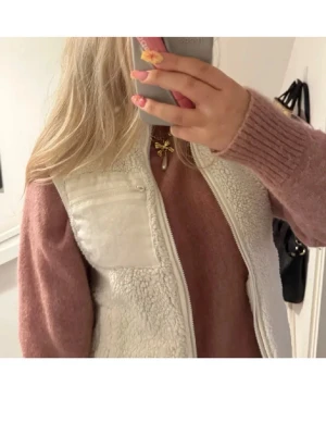 Vit teddyväst - Säljer en vit teddy-dunväst med dragkedja och en bröstficka med dragkedja. Västen har en mjuk, fluffig yta och en stilren look som passar perfekt till lager-på-lager outfits. Perfekt för dig som vill ha en trendig och mysig väst till hösten eller vintern.